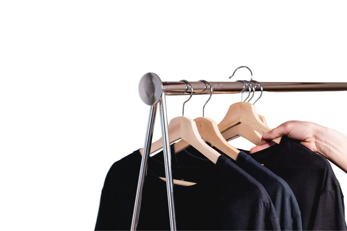 A close-up of a clothing rack with a dark wood horizontal bar, silver metal legs, and wooden hangers holding black crewneck tops. A person's hand and sleeve are visible adjusting the hanger on the right, set against a solid white background.